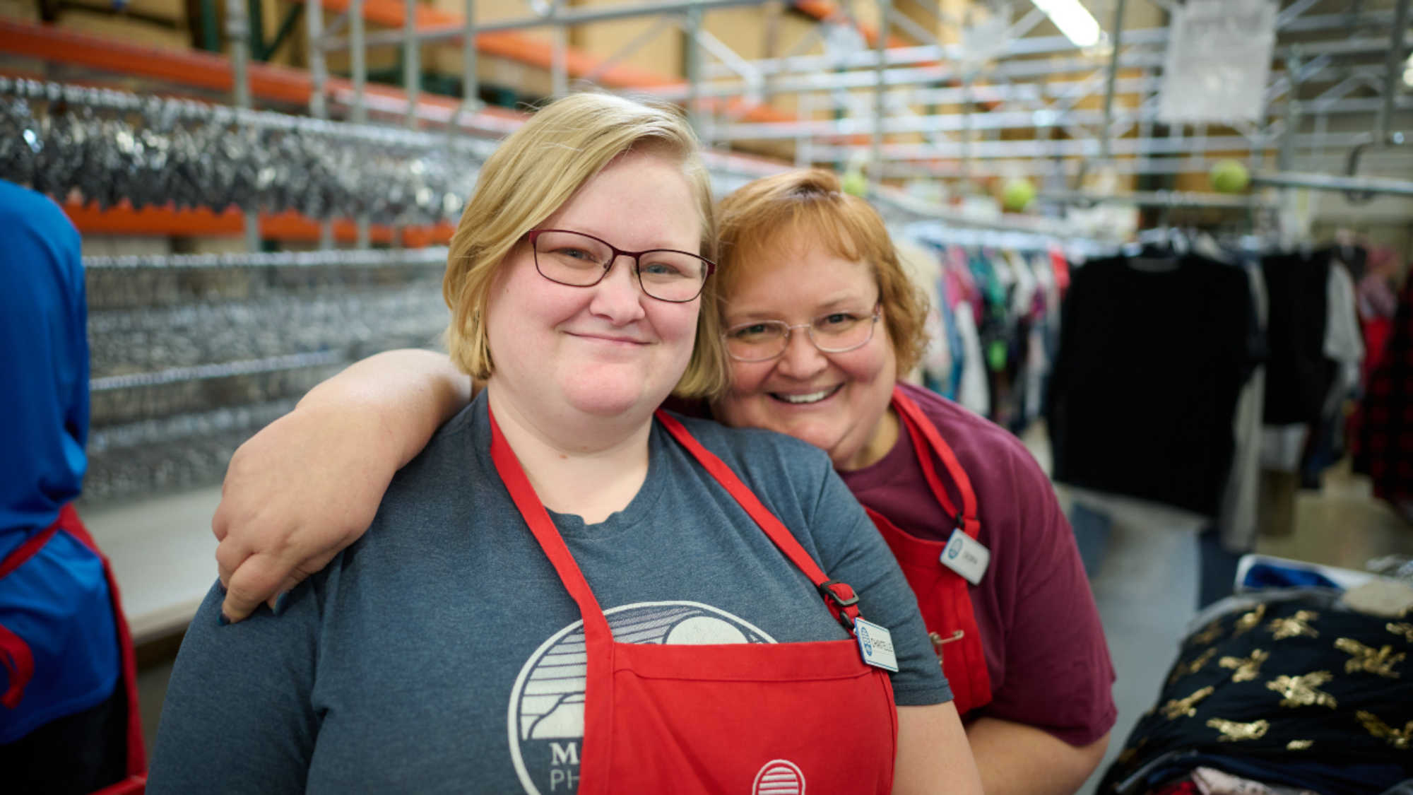 Two female DI associates smile at the camera as they embrace. They are working in the back room, sorting clothing donations.